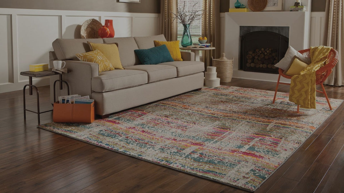A large rectangular area rug with a distressed multicolor pattern in beige, blue, red, and orange tones, styled in a cozy living room next to a beige sofa and a fireplace.