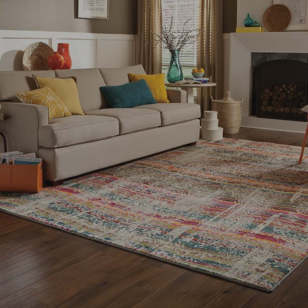 A multicolor abstract patterned area rug with shades of blue, pink, orange, and beige, styled in a living room setting on dark wood floors next to a beige sofa with yellow and teal pillows.