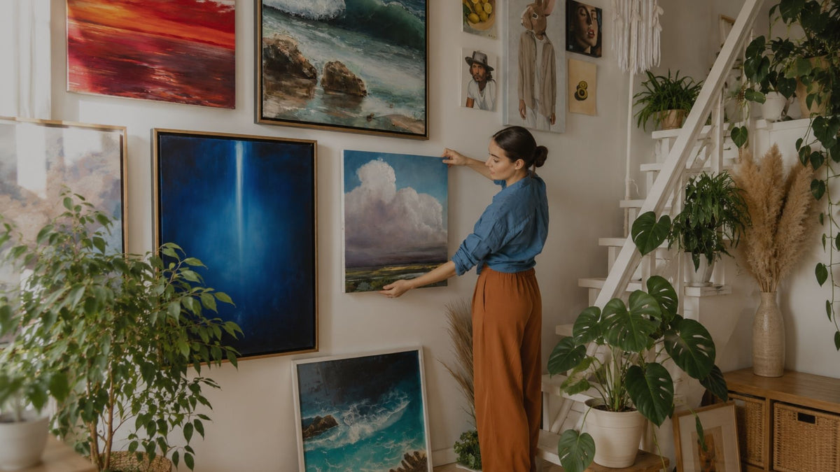 A woman in a blue shirt and rust-orange pants hangs a landscape painting on a wall in a beautifully decorated room with plants, framed art, and a staircase.
